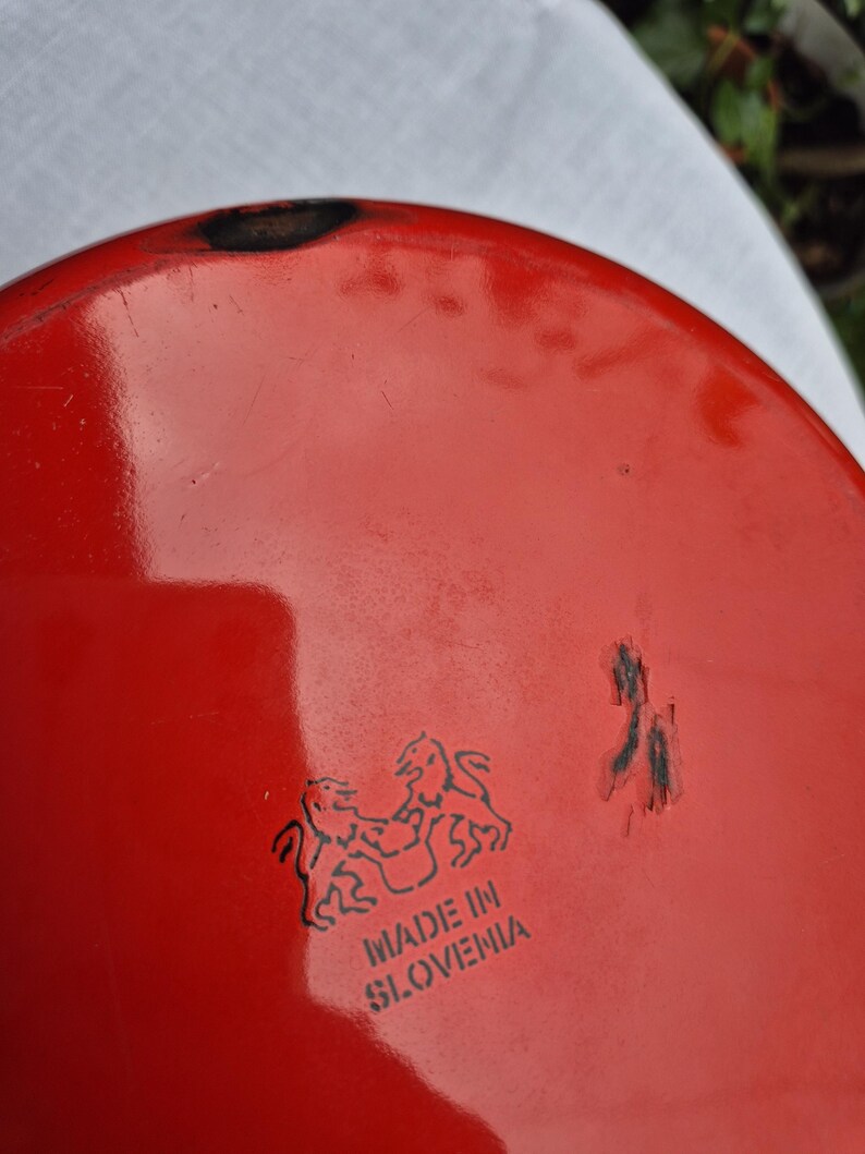 May include: Red enamelware pot with a black rim and a black logo that says "Made in Slovenia" with two lions facing each other.