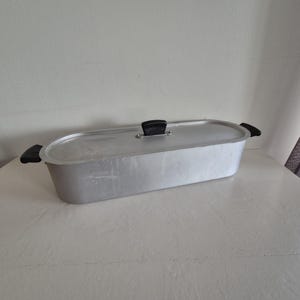 May include: A rectangular, aluminum roasting pan with a lid and black handles. The pan has a black handle on the lid. The pan is sitting on a white surface. Kitchenware.