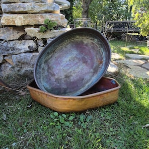 Pode incluir: Duas panelas de metal empilhadas com um acabamento desgastado e oxidado. A panela superior é oval com um interior escuro e manchado, apresentando uma pátina verde e castanha. A panela inferior é um recipiente retangular raso de tom acobreado.