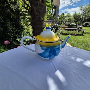 May include: A white, blue, and yellow enamel teapot with a curved handle and spout. The teapot has a blue and white floral design on the body and a yellow lid. The teapot is sitting on a white tablecloth in a garden setting.