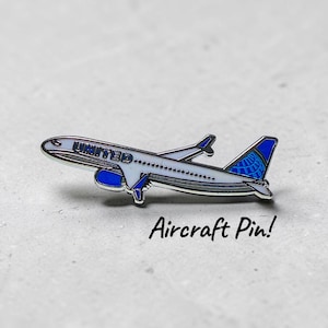 Op de afbeelding: Een zilveren en blauwe vliegtuigspeld met het woord "UNITED" aan de zijkant. De speld heeft een blauwe staart met een wit en blauw logo. De tekst "Aircraft Pin!" staat eronder.