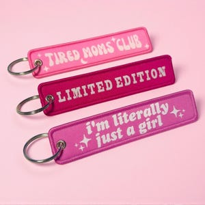 May include: Three embroidered keychains in shades of pink, each with a different white text design. The top keychain reads "TIRED MOMS CLUB", the middle one says "LIMITED EDITION", and the bottom one says "I'm literally just a girl".