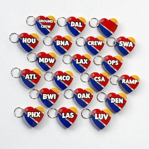 Op de afbeelding: Hartvormige sleutelhangers met een rood, blauw en geel ontwerp. Elke sleutelhanger heeft een driecijferige luchthavencode, waaronder "DAL", "HOU", "LAX" en "LUV". De sleutelhangers zijn gerangschikt op een wit oppervlak.