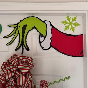 May include: A green hand with black outlines and a red sleeve with white trim, resembling a character from a holiday story, is affixed to a white door. A green snowflake is also attached to the door. A red and white striped bow is at the bottom.