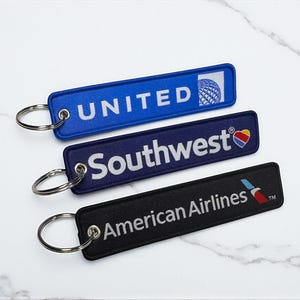 Puede incluir: Tres llaveros rectangulares con texto y logotipos bordados. El superior es azul con "UNITED" y un logotipo de globo. El del medio es azul marino con "Southwest" y un logotipo de corazón. El inferior es negro con "American Airlines".