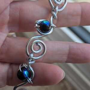 May include: A silver-toned bracelet with a unique, swirling design. The bracelet features black, iridescent beads encased in wire spirals. The beads reflect a spectrum of colors, adding a touch of elegance to the jewelry.