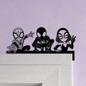 Peut inclure: Une décoration murale en métal noir représentant trois personnages de super-héros stylisés en position accroupie. La figure de gauche a un motif de toile, celle du centre est noire avec un emblème d'araignée, et celle de droite porte une capuche.
