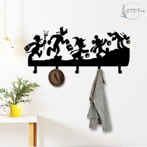 May include: Black metal wall-mounted coat rack featuring silhouettes of cartoon characters in Halloween costumes. The rack has four hooks and is decorated with a hat and a gray garment.
