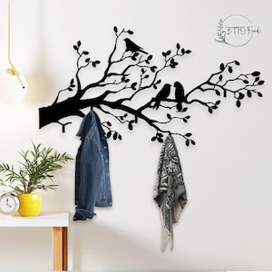 Può includere: Arte da parete in metallo nero raffigurante un ramo d'albero con foglie e uccelli. Il ramo ha ganci per appendere oggetti. Una giacca di jeans e una sciarpa fantasia sono appese ai ganci. L'arte da parete è montata su un muro bianco.