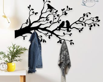 Puede incluir: Arte de pared de metal negro que representa una rama de árbol con hojas y pájaros. La rama tiene ganchos para colgar objetos. Una chaqueta vaquera y una bufanda estampada cuelgan de los ganchos. El arte de pared está montado en una pared blanca.