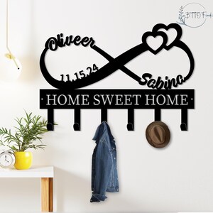 May include: Black metal wall-mounted coat rack with an infinity symbol design. The rack features the names "Oliveer" and "Sabina", the date "11.15.24", and the phrase "HOME SWEET HOME". A denim jacket and a brown hat hang from the hooks.