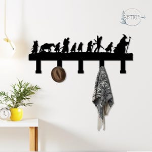 Könnte beinhalten: Schwarzer Wand-Kleiderhaken aus Metall mit einer Silhouette einer Figurengruppe. Ein brauner Hut und ein gemusterter Schal hängen an den Haken. Der Haken ist an einer weißen Wand befestigt. Ein kleiner Tisch und eine Pflanze sind sichtbar.