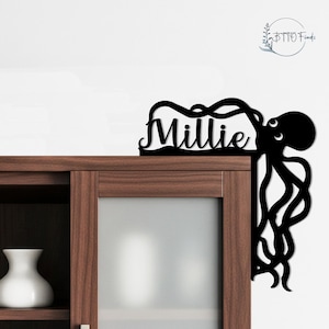May include: Black metal octopus wall art with the name "Millie" cut out. The octopus design is placed on the corner of a wooden cabinet. The tentacles extend down the side. The name is in a cursive font.