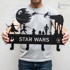 May include: Black metal Star Wars key holder featuring iconic characters and symbols. The design includes the Death Star, X-wing fighters, and an AT-AT walker. The text "STAR WARS" is prominently displayed. The key holder has four hooks for keys.