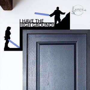 Könnte beinhalten: Schwarze Metall-Wandkunst mit zwei silhouettierten Figuren mit blauen Lichtschwertern. Der Text "I HAVE THE HIGH GROUND!" ist zu sehen. Die Kunst ist für die Wand- oder Türmontage konzipiert und bietet ein dekoratives Element im Science-Fiction-Stil.