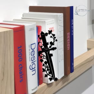 Könnte beinhalten: Ein schwarzer metallener, schwertförmiger Lesezeichen mit floralen Akzenten steht zwischen Büchern mit weißen, roten und braunen Einbänden. Die Bücher zeigen Titel wie 'Design' und '1000 Chairs'. Das Lesezeichen verleiht dem Bücherregal eine dekorative Note.
