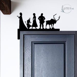 May include: A black metal door topper featuring silhouettes of characters from the animated film "Frozen." The design includes figures of Elsa, Olaf, Anna, Kristoff, and Sven. The topper is mounted on a dark blue door.