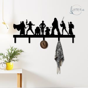 May include: Black metal wall-mounted coat rack featuring silhouettes of Star Wars characters. The rack has several hooks for hanging items, with a hat and scarf hanging from the hooks. The wall is white, and the rack is mounted on it.