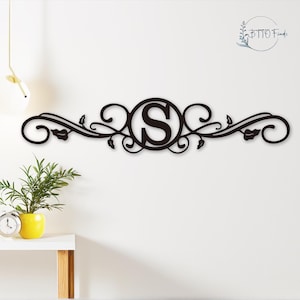 Op de afbeelding: Zwarte metalen wanddecoratie met een gerold ontwerp en een cirkel in het midden met de letter "S".