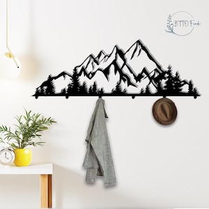 Può includere: Appendiabiti da parete in metallo nero con un design a silhouette di montagna e foresta. Il portabiti ha diversi ganci per appendere cappotti, cappelli o altri oggetti. Una giacca grigia e un cappello marrone sono appesi ai ganci.