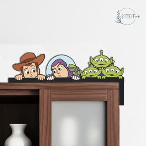 Op de afbeelding: Een decoratieve houten plankopzet met personages uit de animatiefilm "Toy Story". Het ontwerp omvat Woody, Buzz Lightyear en drie Little Green Men, allemaal over een zwarte richel kijkend. De plank is gemonteerd op een houten kast.