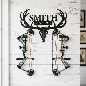 May include: Black metal bow rack with the name "SMITH" and "EST.1998" cut out. The rack features a deer skull design and holds two bows, one green and black, the other tan and black, against a white wood-paneled wall.