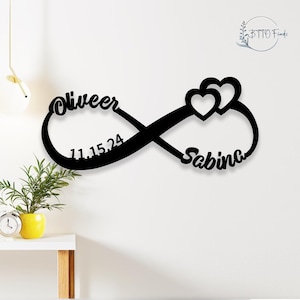 May include: Black metal infinity symbol with the names "Oliver" and "Sabina" and the date "11.15.24" inscribed on it. Two hearts are intertwined in the centre of the symbol.