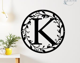 Peut inclure: Décoration murale en métal noir avec la lettre "K" entourée d'une guirlande de feuilles. L'œuvre est fixée sur un mur blanc, créant un contraste saisissant. Le design comprend le texte "BTTO Finds".