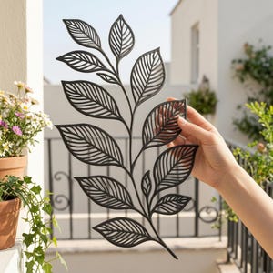 Op de afbeelding: Zwarte metalen blad tak wanddecoratie met gedetailleerde bladeren uitsparingen. Het decoratieve stuk wordt door een hand vastgehouden, wat het ingewikkelde ontwerp laat zien. De achtergrond toont een balkon en potplanten.