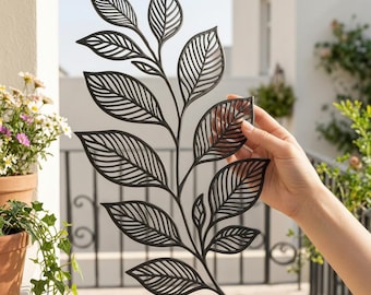 May include: A black metal leaf branch wall decor with detailed leaf cutouts. The decorative piece is held by a hand, showcasing its intricate design. The background features a balcony and potted plants, suggesting an outdoor or garden theme.
