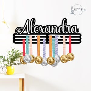 May include: A black wooden medal display with the name "Alexandra" in white script. The display holds several medals with colorful ribbons. The medals are gold and silver in color. The display is mounted on a white wall.