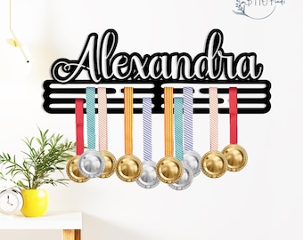 May include: A black wooden medal display with the name "Alexandra" in white script. The display holds several medals with colourful ribbons. The medals are gold and silver in colour. The display is mounted on a white wall.