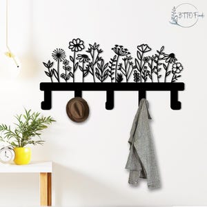 May include: Black metal wall-mounted coat rack featuring a floral design. The rack has four hooks and is decorated with cut-out flowers and foliage. A brown hat and a gray scarf hang from the hooks. The Etsy shop logo "BTTO Finds" is in the upper right corner.