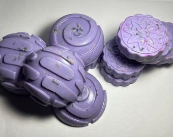 May include: A set of six lavender-coloured soap bars with different designs. The soaps are round, square, and circular with a floral design. The soaps are made with natural ingredients and are perfect for a relaxing bath.
