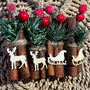 May include: Four cinnamon sticks decorated with red berries, green pine sprigs, and wooden reindeer and sleigh ornaments.