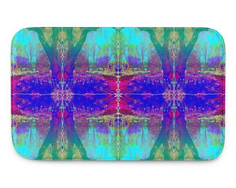 May include: Rectangular bath mat with a vibrant, symmetrical design. The artwork features a mirrored landscape with trees, water, and foliage in shades of blue, purple, green, and pink. The mat has rounded corners and a green border.