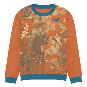 May include: A long-sleeve crewneck sweater in burnt orange with teal trim at the collar, cuffs, and hem. The front features a detailed, textured graphic print in shades of brown, cream, and teal, depicting a profile silhouette and a butterfly.