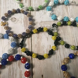 May include: A collection of beaded bracelets in various colors, including green, blue, yellow, red, and gold. The bracelets feature sparkling, round beads strung together on a chain. The bracelets are displayed on a light-colored wooden surface.