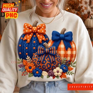 May include: A cream-colored sweatshirt featuring a graphic of three pumpkins with knitted textures. The left pumpkin is blue with orange polka dots and an orange bow. The right is orange with a blue plaid pattern and blue bow. The center is leopard print.