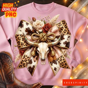 May include: Pale pink sweatshirt with a cow skull design, bow, and floral accents. The design incorporates leopard print, roses, feathers, and a gold rope. The text "COCOSPIRITY" is visible in the bottom right corner of the image.