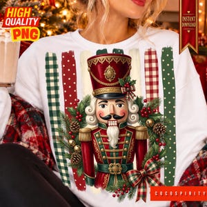 May include: A white sweatshirt featuring a graphic of a nutcracker in a red and green uniform, surrounded by a wreath of pine branches, red berries, and pinecones. The background includes vertical stripes of green plaid, red dots, and red gingham.