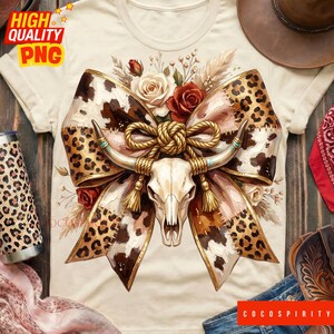 May include: A cream-coloured t-shirt with a detailed graphic. It features a cow skull with long horns, a large bow with a cowhide and leopard print design, and floral accents. The shirt has the text "HIGH QUALITY PNG" in the upper left corner.