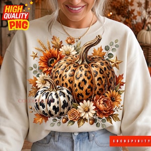 May include: A cream-colored sweatshirt featuring a graphic print of two pumpkins with leopard and cow print patterns, surrounded by sunflowers, daisies, autumn leaves, wheat stalks, and gold stars. The text COCOSPIRITY is printed at the bottom.