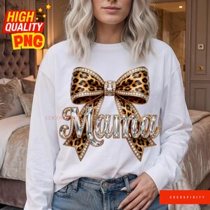 May include: White long-sleeve shirt with a large bow design. The bow features a leopard print pattern and is embellished with rhinestones. The word "Mama" is written in silver, rhinestone-studded letters.