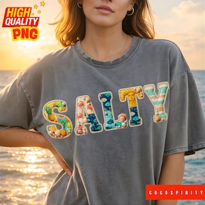 May include: Gray t-shirt with the word "SALTY" in colorful, textured letters. The letters are decorated with images of fruits, crabs, fish, seashells, and a sun. The shirt is worn by a person near the ocean.