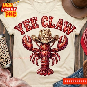 Könnte beinhalten: Cremefarbenes T-Shirt mit einem roten Hummer, der einen Cowboyhut trägt. Der Text "YEE CLAW" steht in einer roten, mit Edelsteinen besetzten Schriftart über dem Hummer. Das Design kombiniert Country- und Meeresfrüchte-Themen.