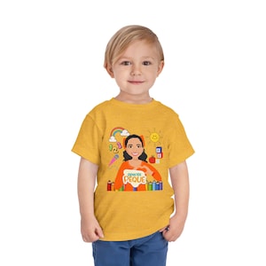 May include: A toddler wearing a mustard yellow t-shirt with a colorful graphic. The graphic features a cartoon woman with the text "APRENDE PEQUE" and educational elements like a rainbow, numbers, and crayons.