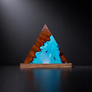 May include: A triangular wooden lamp base with a blue resin insert depicting a scene of underwater life. The resin is translucent and glows from within, creating a luminous effect.