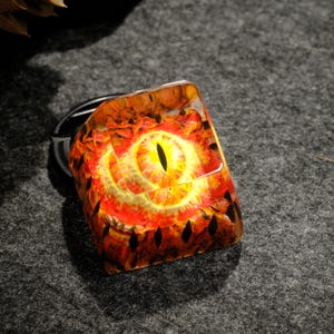 Puede incluir: Un tapón de tecla de resina con un diseño de ojo rojo y amarillo. El ojo está rodeado de un patrón negro y naranja.