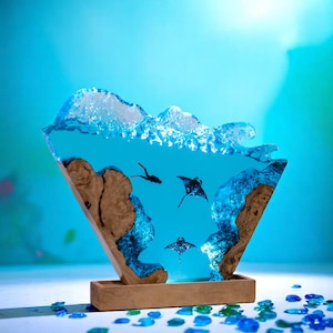 May include: A unique resin sculpture depicting an underwater scene. The artwork features a diver, manta rays, and a wave cresting above. The sculpture is set in a wooden base, with blue and brown tones.